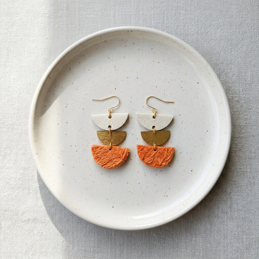 A pair of handmade semi-circular dangle earrings with gold plated hooks, featuring a white leather top half, a textured brass middle half, and an orange cotton bottom half, displayed on a light gray background.