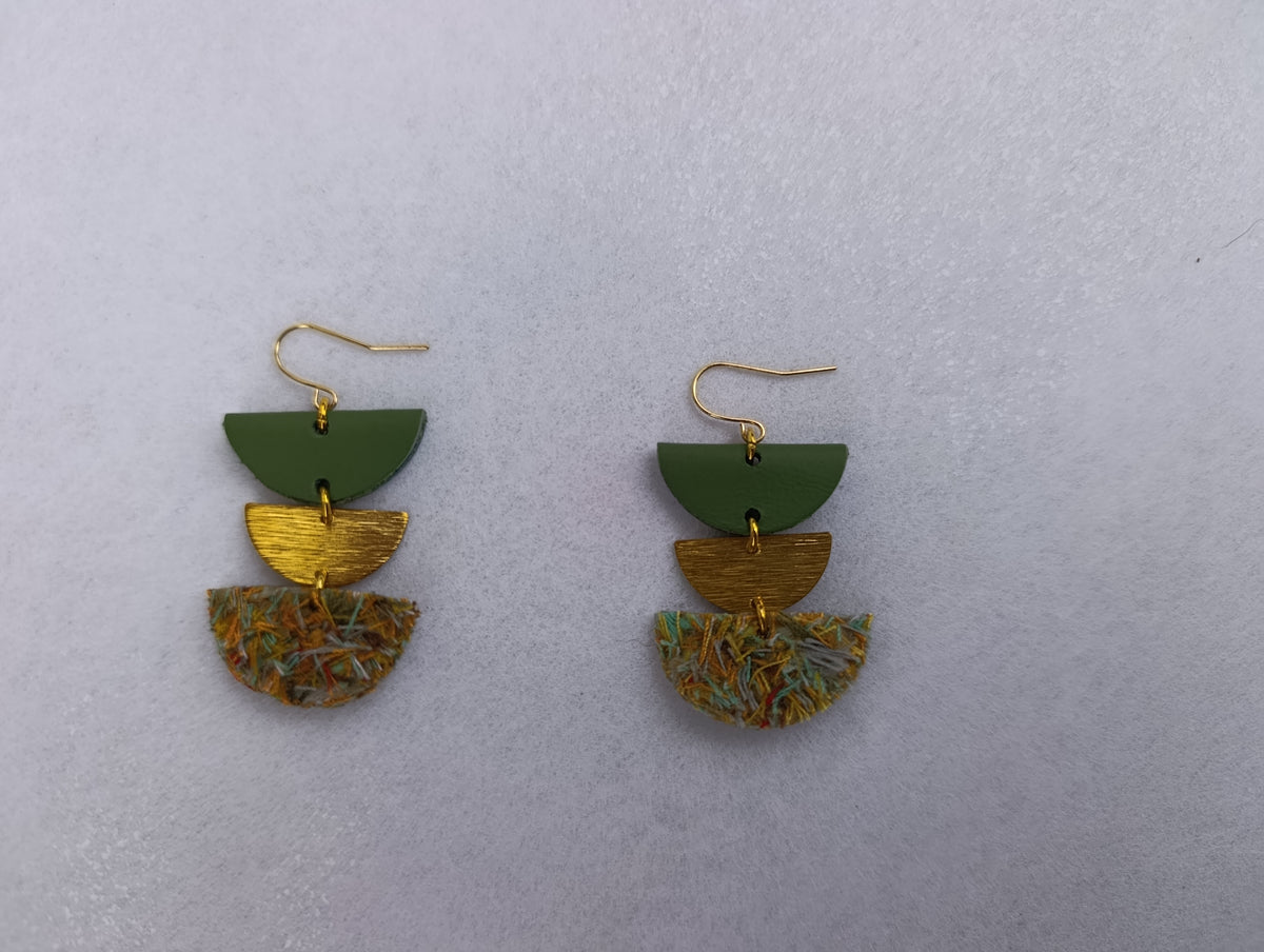 A pair of handmade geometric dangle earrings on a textured light gray background. Each earring features a gold-tone fishhook wire, a dark green leather semi-circle, a textured gold semi-circle, and a textured multicolor semi-circle made of shredded cotton.