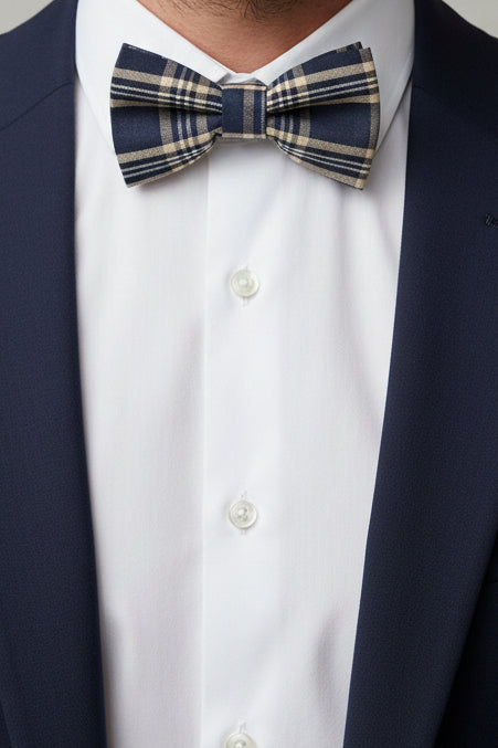 A classic plaid bow tie in navy blue, beige, and white, laid flat on a white background with faint paint splatters.